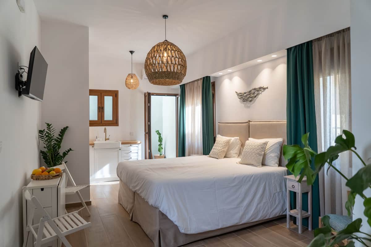 A modern bedroom with a large bed, bedside tables, a TV, and a kitchen area in the background.