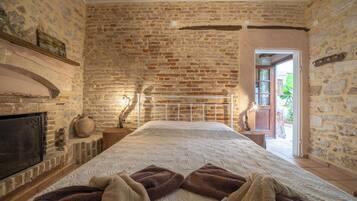 A bedroom with a brick wall, a fireplace, a bed with a white and brown patterned bedspread, and a view of the outside through a door.