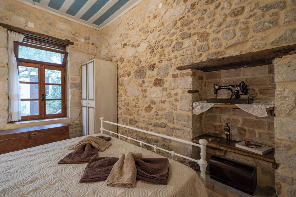 A stone-walled room with a bed, a window with curtains, a wooden dresser, and a fireplace with a shelf above it.