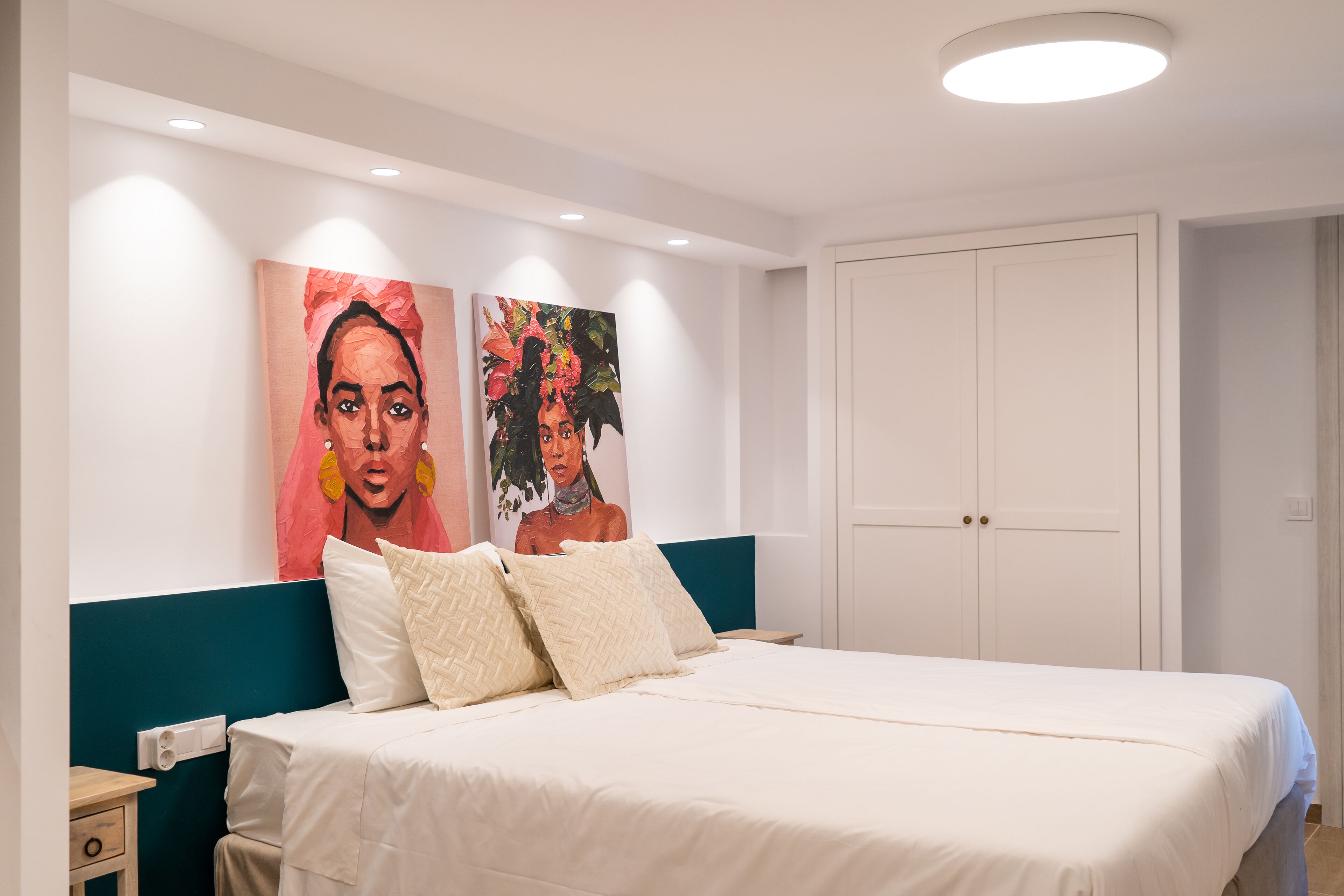 A bedroom with a bed, two paintings on the wall, a nightstand, and a wardrobe.