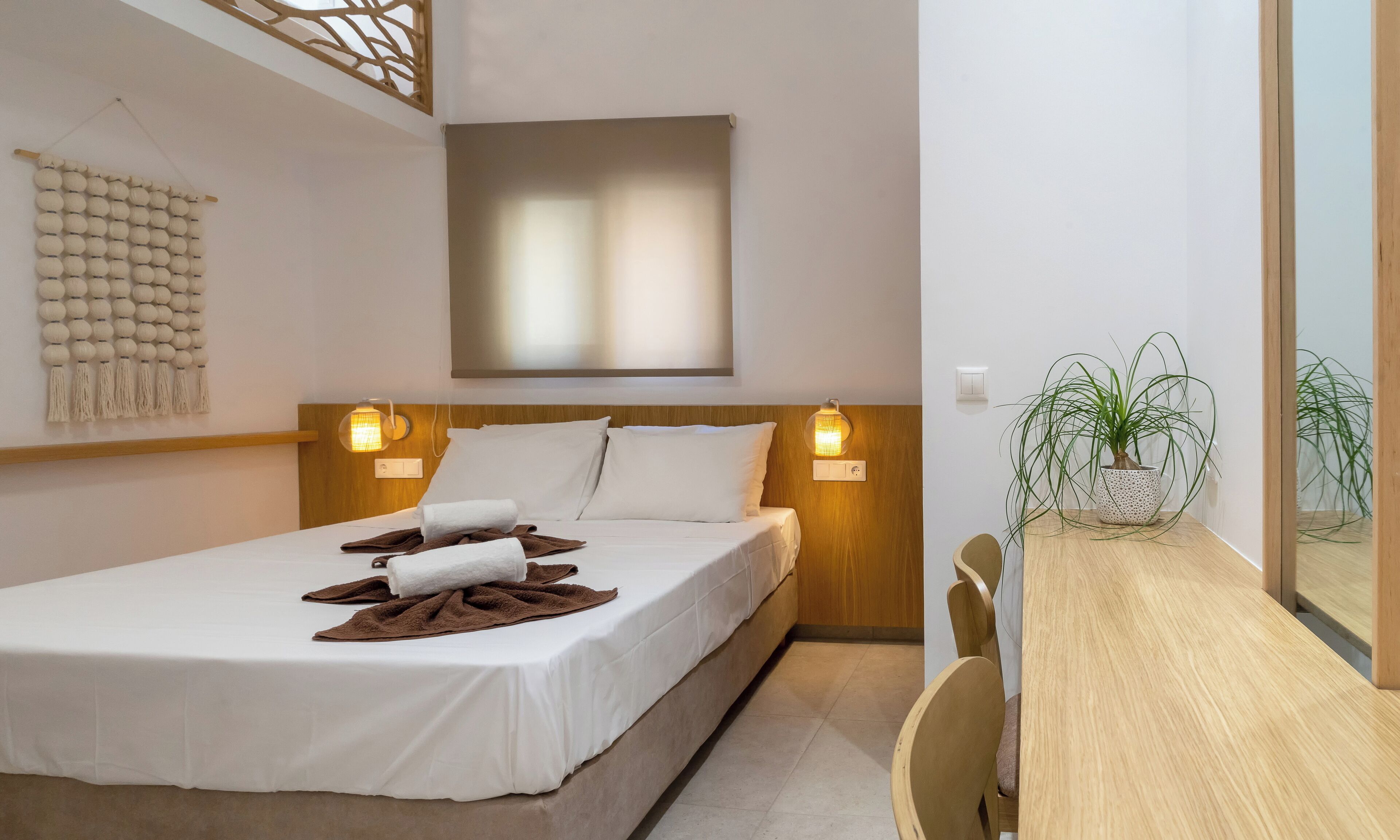 A neatly made bed with white linens and brown towels, a wooden headboard, and two wall-mounted lamps.