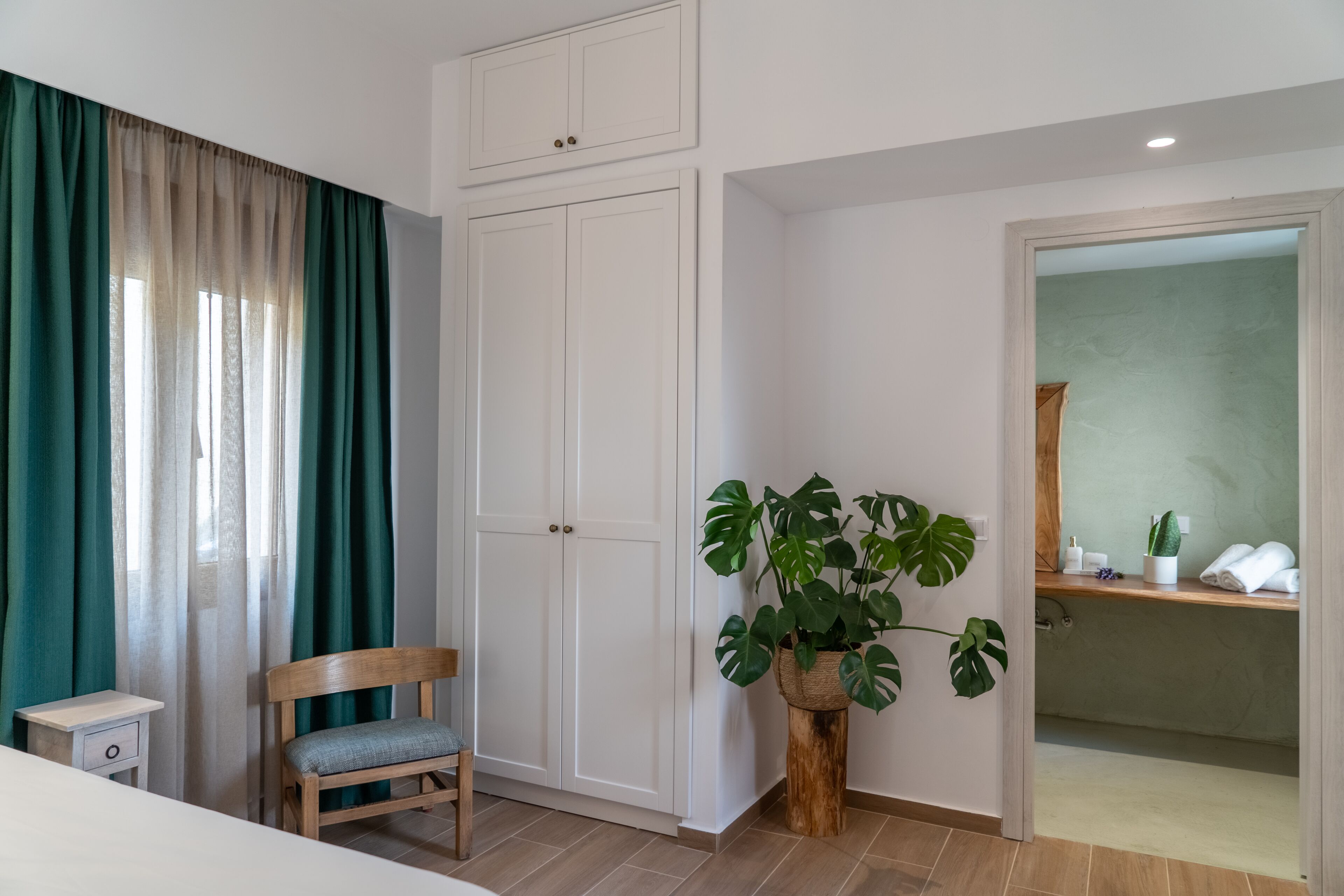 Ein Schlafzimmer mit einem Bett, einem Holzstuhl, einem Kleiderschrank, einer Pflanze und einem durch eine offene Tür einsehbaren Badezimmer.