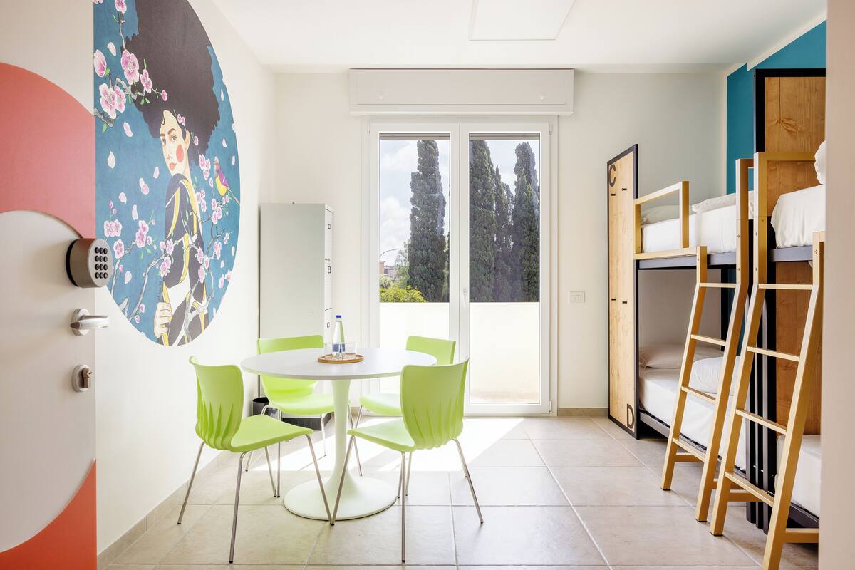 A compact room with a dining table and chairs, a bunk bed, and a large window with a view of trees.