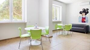 A bright, modern room with a round dining table, green chairs, a gray sofa, and large windows offering a view of trees.
