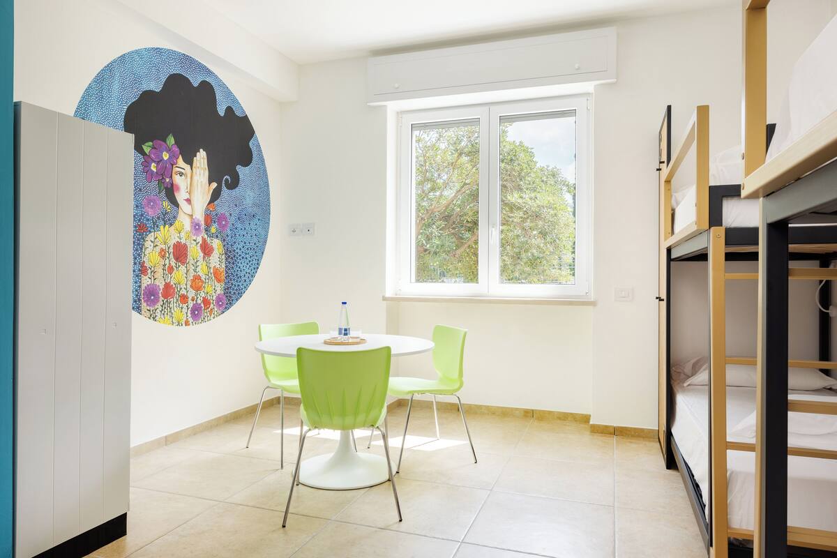 A room with a round table, green chairs, bunk beds, a window, and a wall mural.