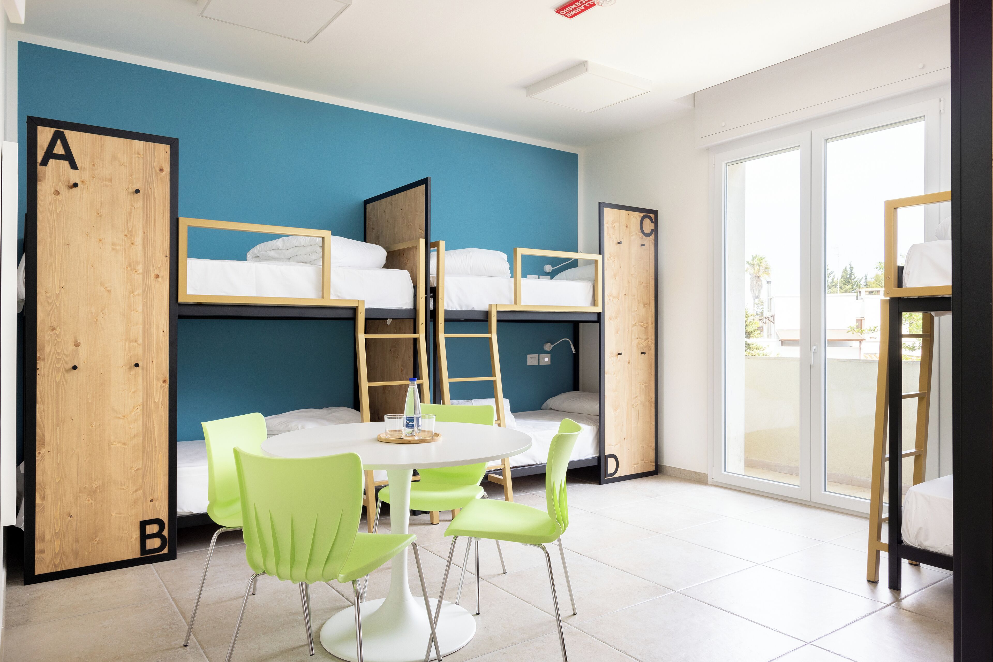 A dormitory room with bunk beds, a round table, and green chairs.