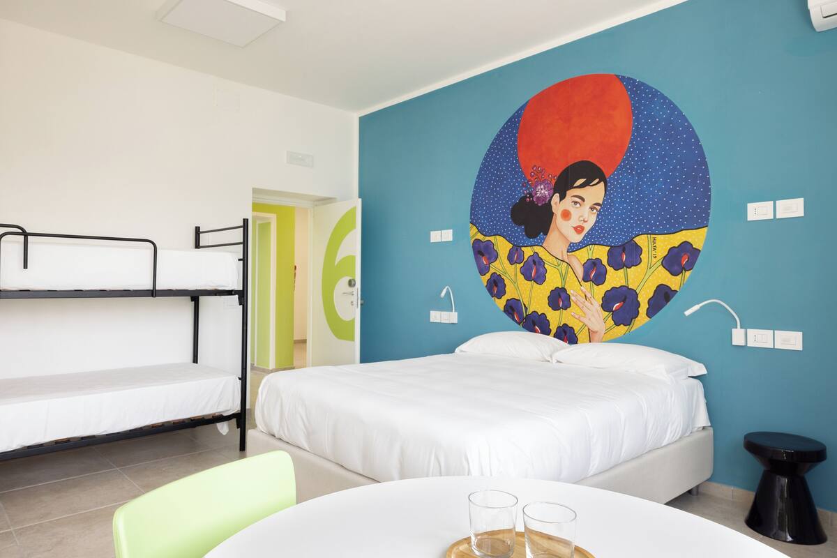 A room with a bunk bed, a single bed, a round table with glasses, and a colorful mural on the wall.