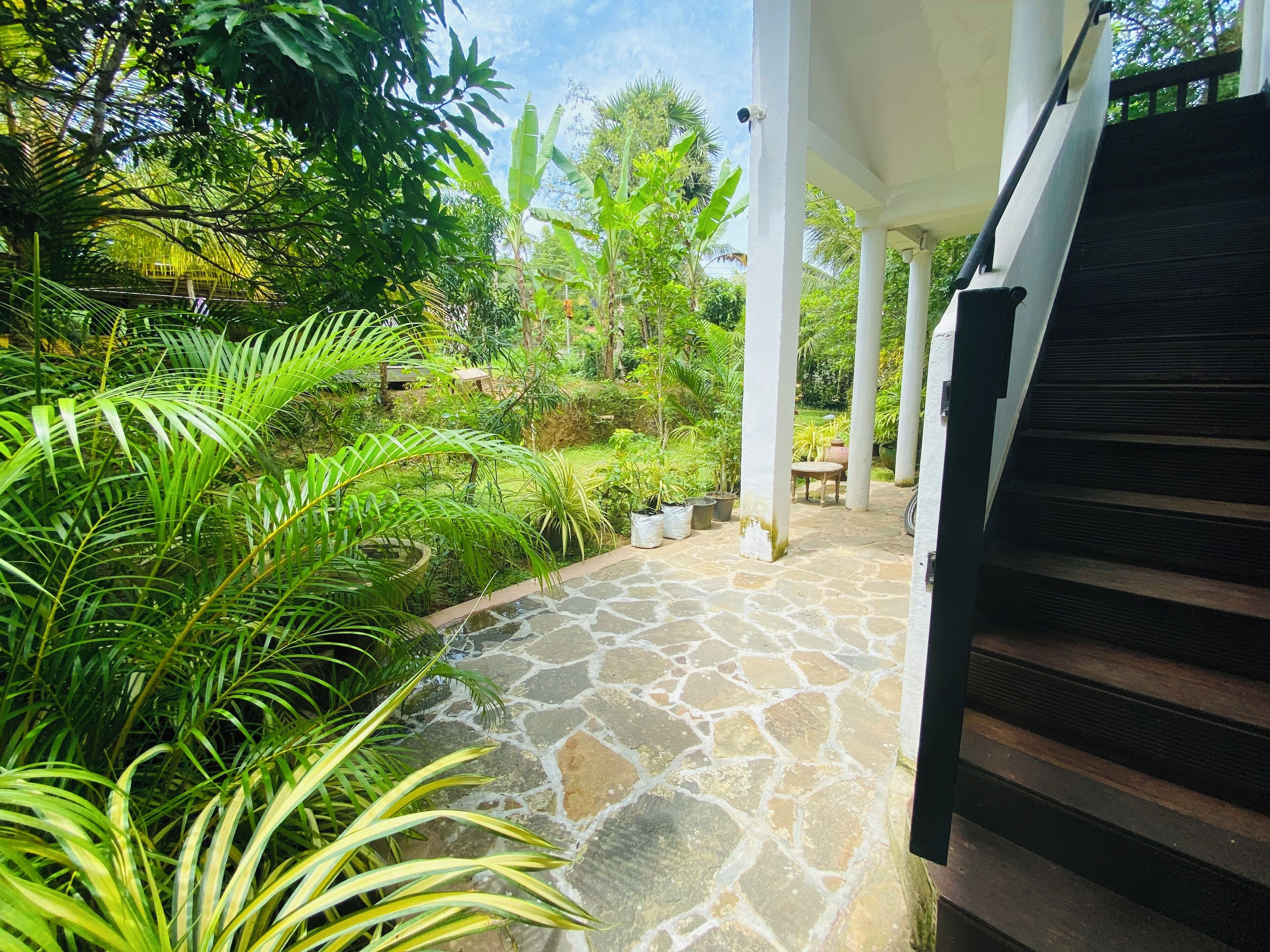 A stone pathway leading to a covered area with a table and chairs, surrounded by lush greenery and a security camera.