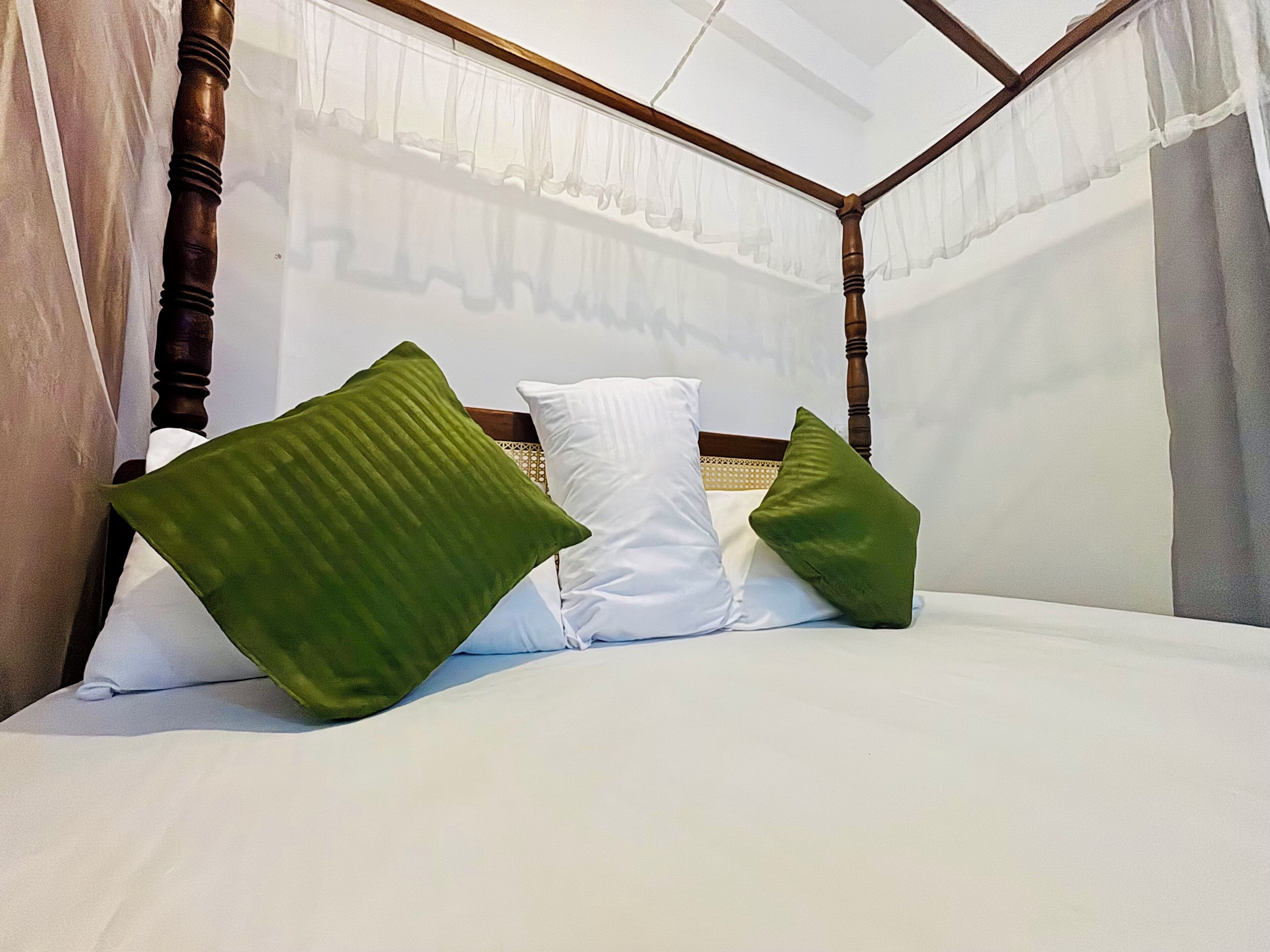 A bed with white bedding and green pillows, a wooden headboard, and sheer curtains.