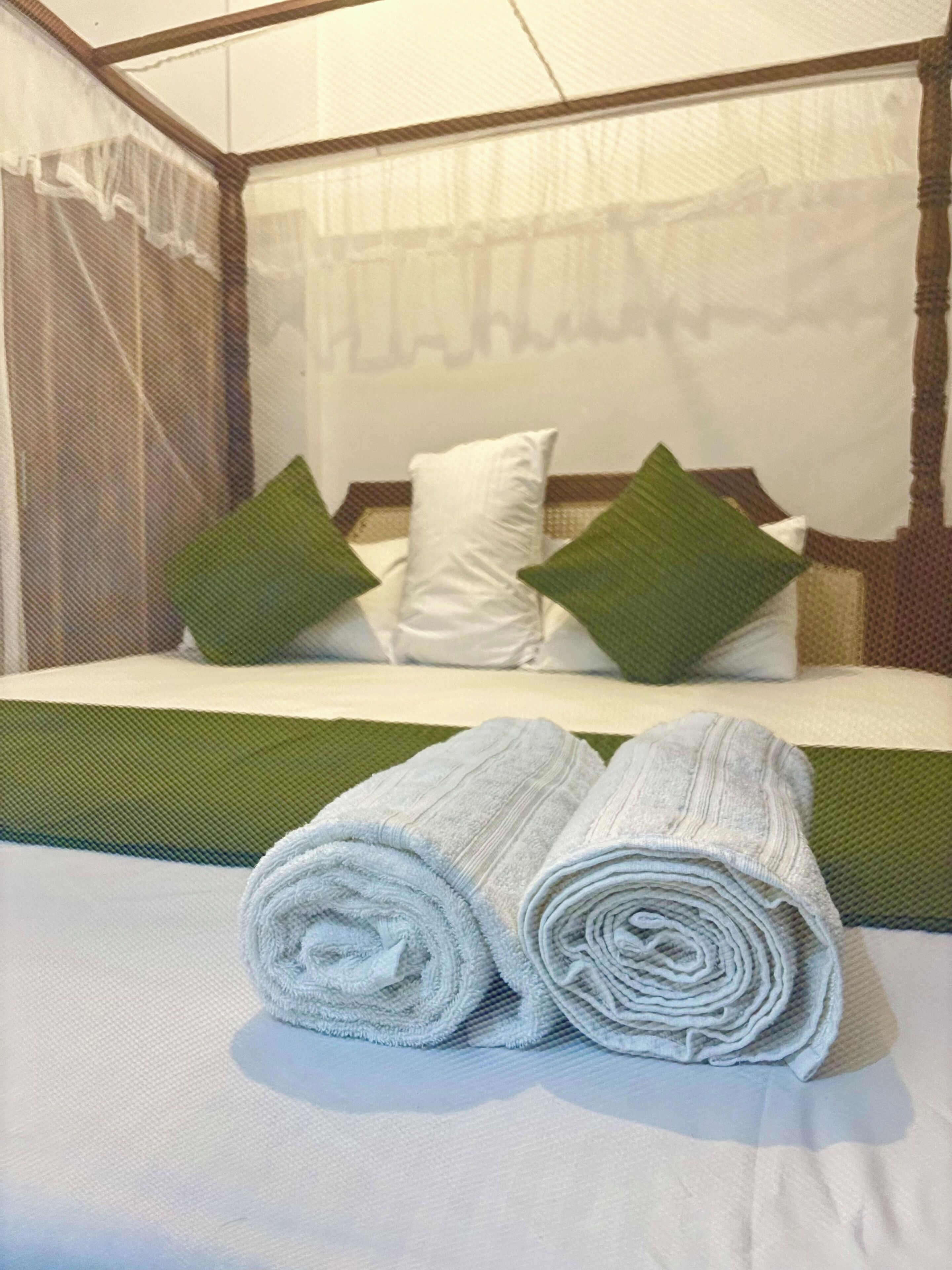 A neatly made bed with green and white pillows, a white headboard, and two rolled towels on the bed.