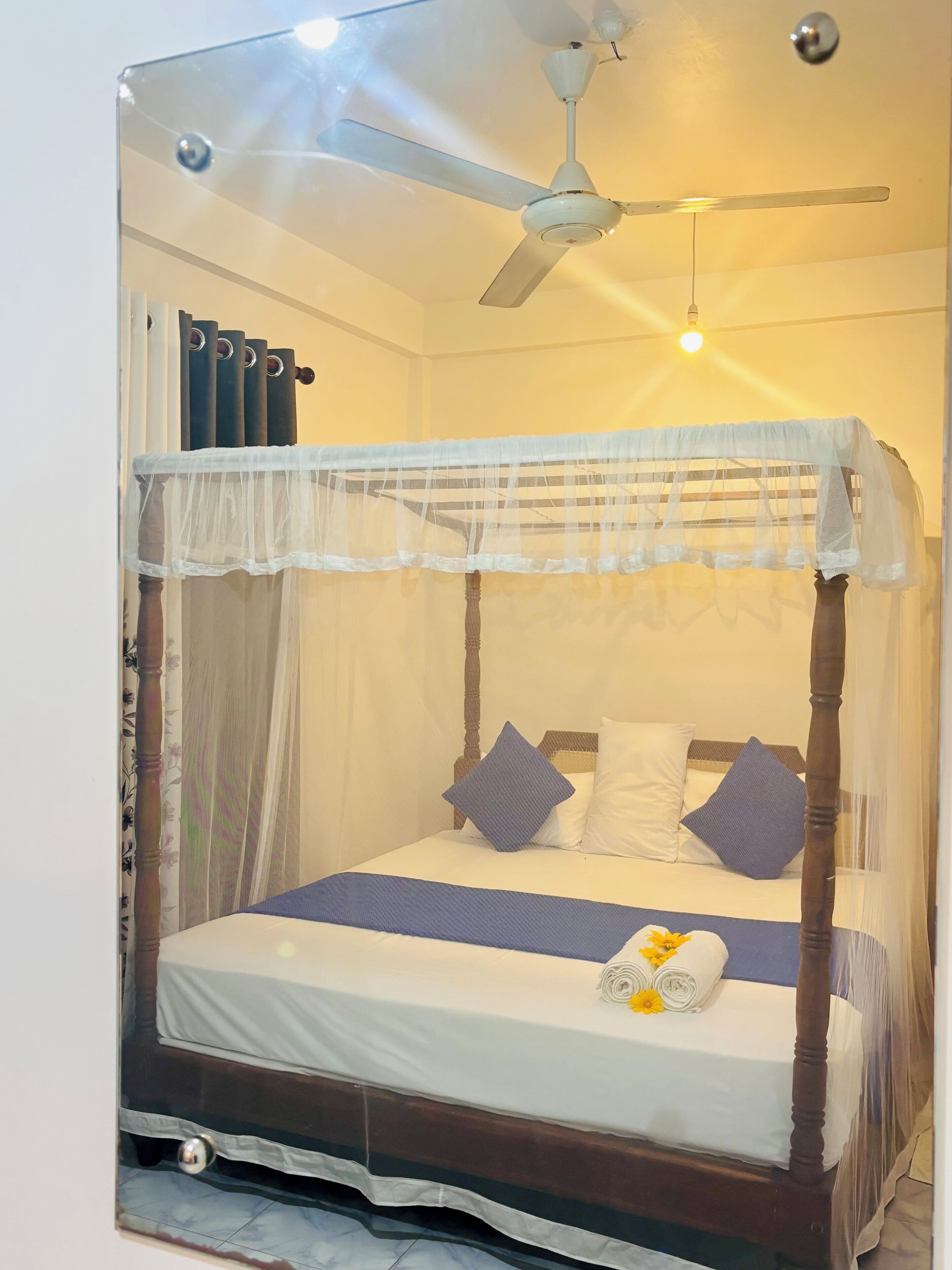 Ein Himmelbett mit Baldachin, ein Deckenventilator und ein Nachttisch mit gerolltem Handtuch und Blumen.