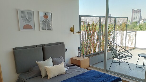 Un dormitorio moderno con una cama grande, una mesita de noche, una silla y un balcón con vistas a edificios.
