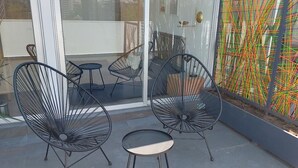 Un espacio exterior moderno con sillas de metal y una pequeña mesa, una puerta de cristal que da a un balcón y una llamativa obra de arte abstracta en la pared.