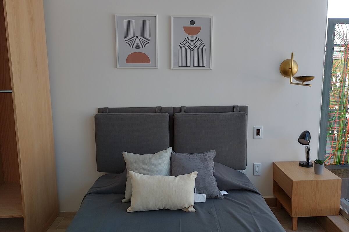 A modern bedroom with a grey bed, two pillows, a bedside table with a lamp, and two abstract wall art pieces.