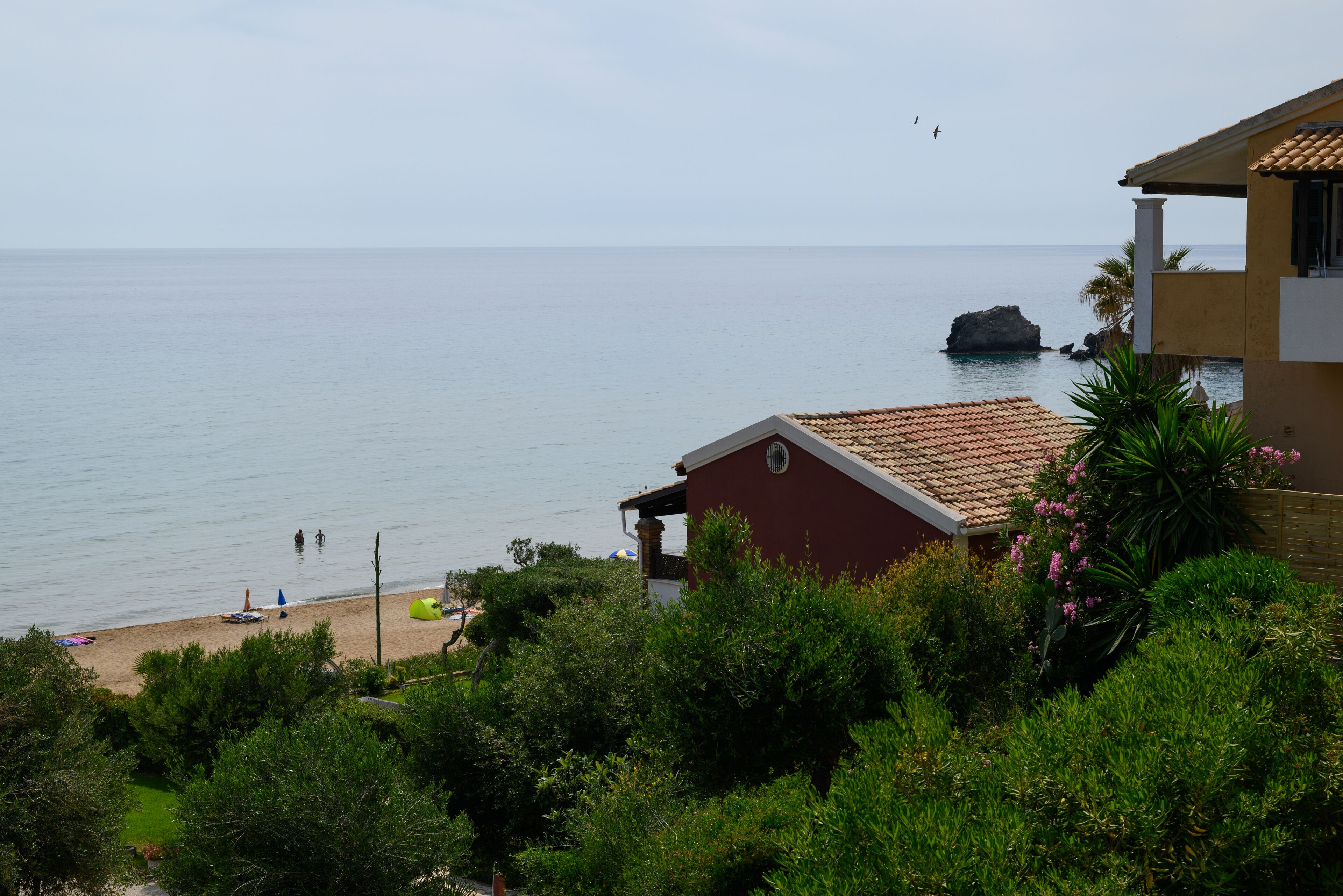 A coastal view with a beach, a red building, and a rocky outcrop in the sea.