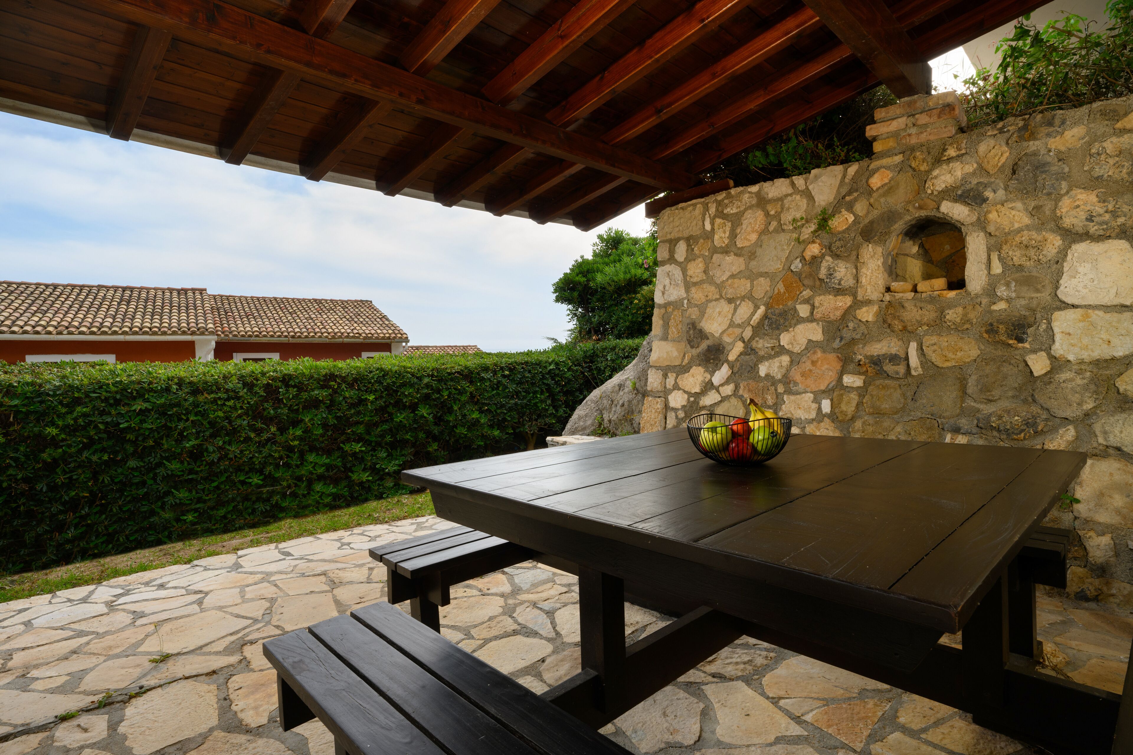 A stone patio with a wooden table and benches, a stone wall with a window, and a building with a tiled roof in the background.