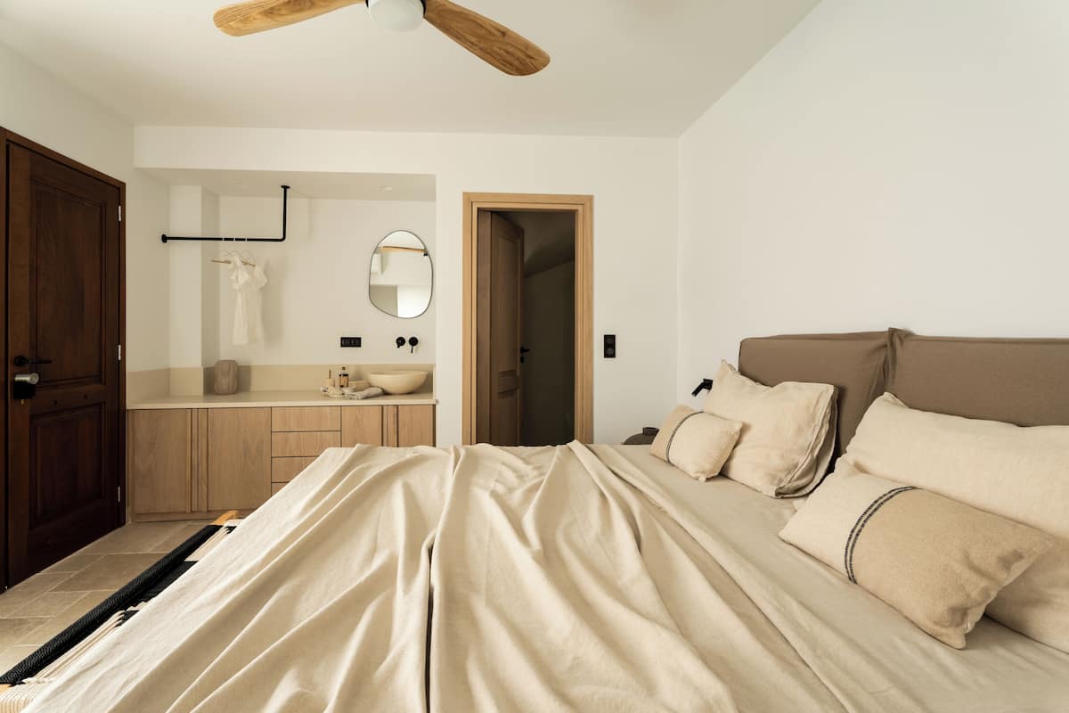 A bedroom with a bed, a wooden door, a bathroom with a mirror, and a ceiling fan.