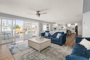 A spacious living room with a large sliding glass door leading to a balcony, a ceiling fan, and a kitchen area in the background. - Five Bedroom Waterfront Home with Lots of Space (Ocean City)