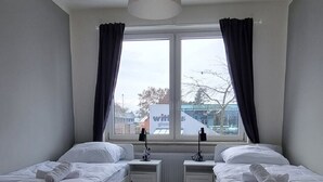 Et soveværelse med to senge, en ventilator i loftet, et vindue med gardiner og udsigt ud.