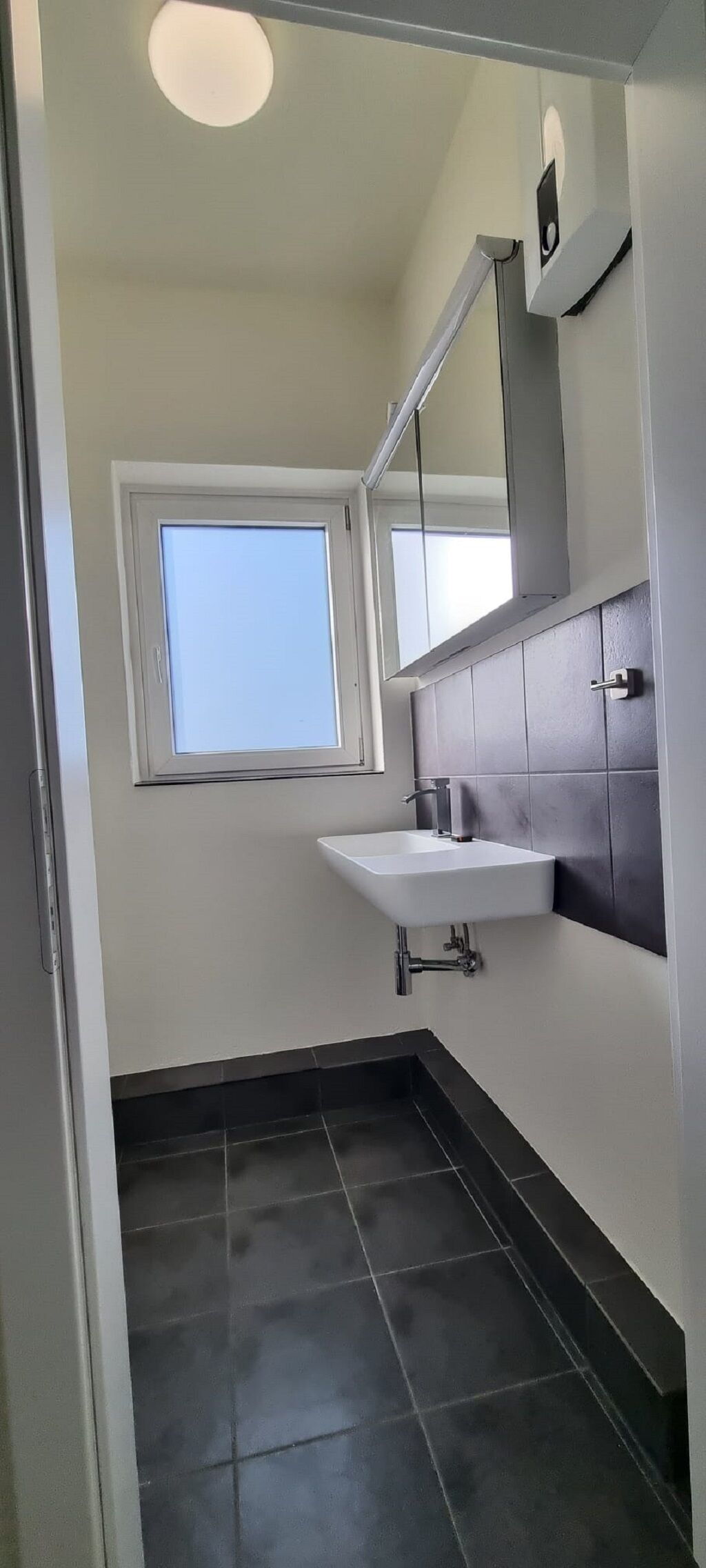 A small bathroom with a window, a white sink, a mirror, and dark tiled flooring.