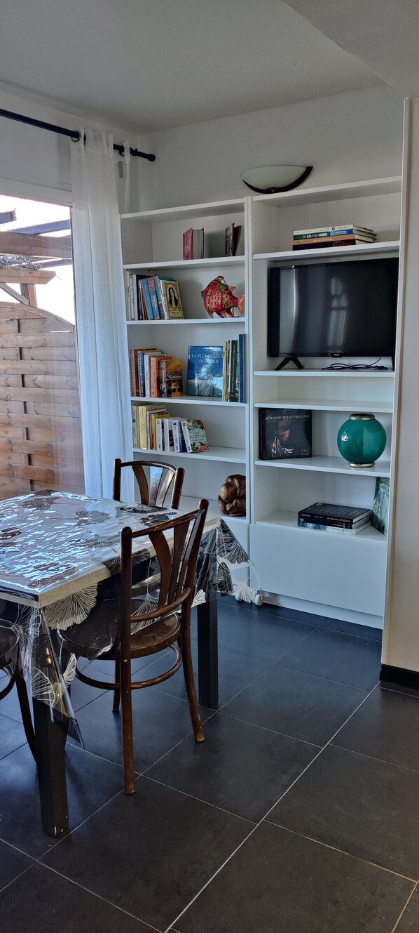 A dining area with a table covered in a patterned tablecloth, two wooden chairs, and a bookshelf with books and decorative items. - Emplacement n1, F2 Bord de Plage et Départ du Sentier des Douaniers (ROGLIANO)