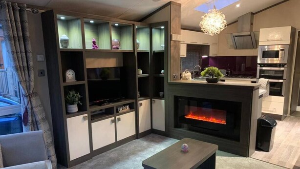 A modern living room with a fireplace, built-in shelves, and a kitchen area with appliances. - Stunning 3-bed Lodge in Windermere (Windermere)