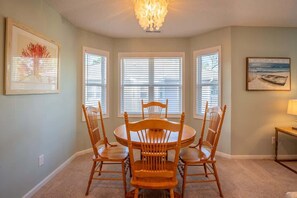 A dining area with a round table and four chairs, large windows with blinds, and artwork on the walls.