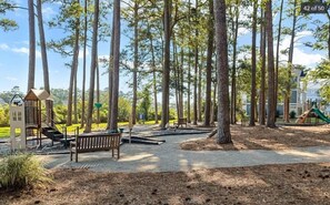 A park with a playground, a wooden bench, and tall trees.