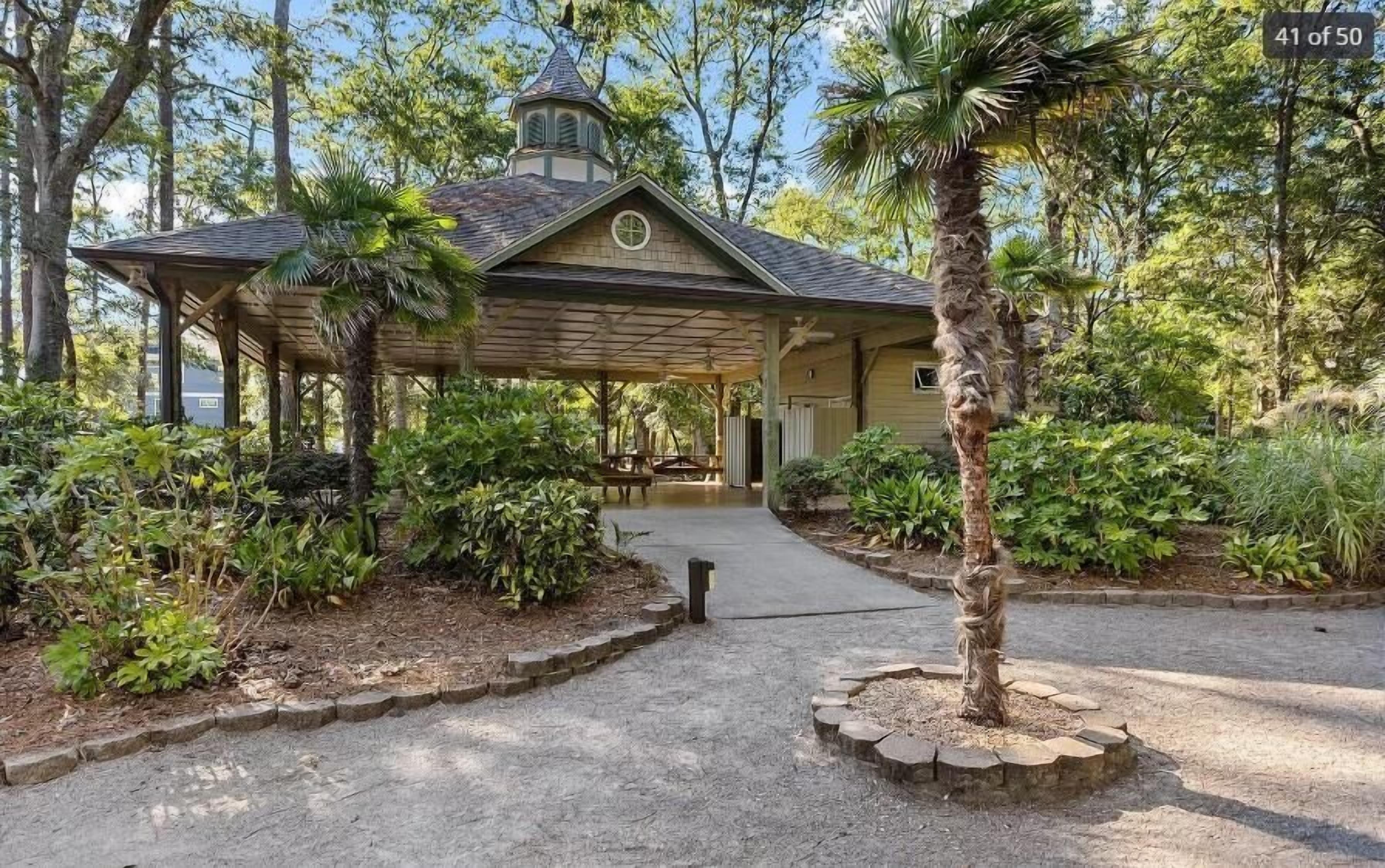 A covered pavilion with a roof, surrounded by lush greenery and a paved path leading to it.