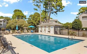 Una piscina rectangular con sillones de descanso y sombrillas, rodeada de árboles y un pequeño edificio con techo.