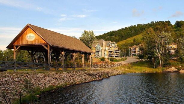 A lodge with a wooden pavilion, a lake, and a hillside with houses. - Ski-In/Ski-Out Mont-Blanc Condo w/ Pool & Lake Access (Mont-Blanc)