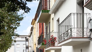 Una calle estrecha flanqueada por edificios, con balcones de barandilla metálica y macetas con flores.