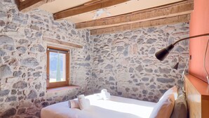 Ein Schlafzimmer mit Steinwänden, Holzbalkendecke, einem Bett mit weißer Bettwäsche, einem kleinen Fenster und einer Nachttischlampe.