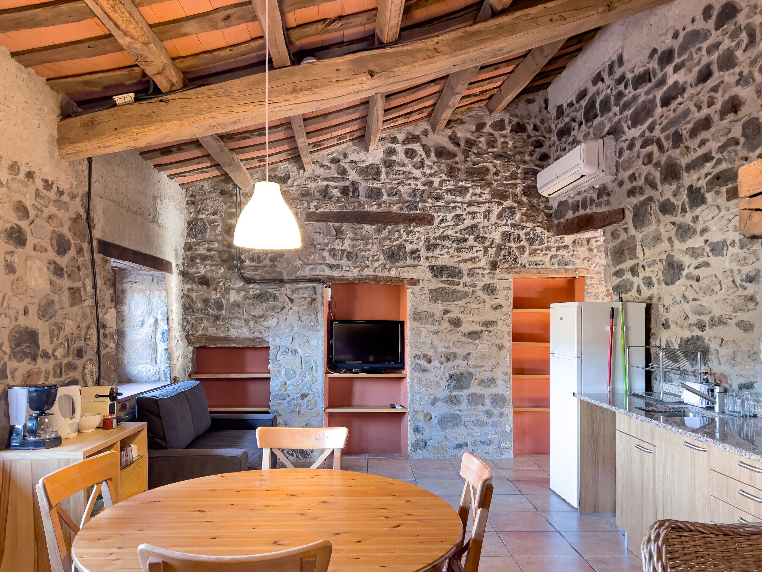 A rustic interior with a stone wall, wooden ceiling beams, a round wooden table, and a television in a stone alcove.