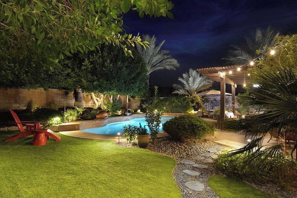 Un espace extérieur aménagé pour la nuit, comprenant une piscine, un coin salon couvert et un chemin en pierre.