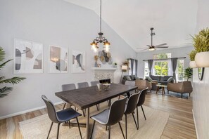 A dining area with a long table, chairs, and a chandelier. There is a fireplace, a sofa, and a ceiling fan in the background. - Private Saltwater Pool & Hot Tub Oasis w/ WiFi (La Quinta)