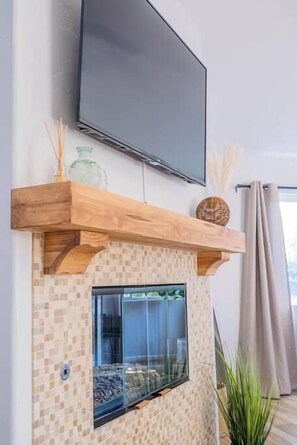 Un salon moderne avec un téléviseur à écran plat fixé au mur, une étagère en bois ornée d’objets décoratifs, une cheminée en verre et une plante en pot.