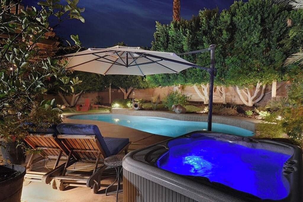 A hot tub with a blue light, a pool, lounge chairs, and an umbrella.