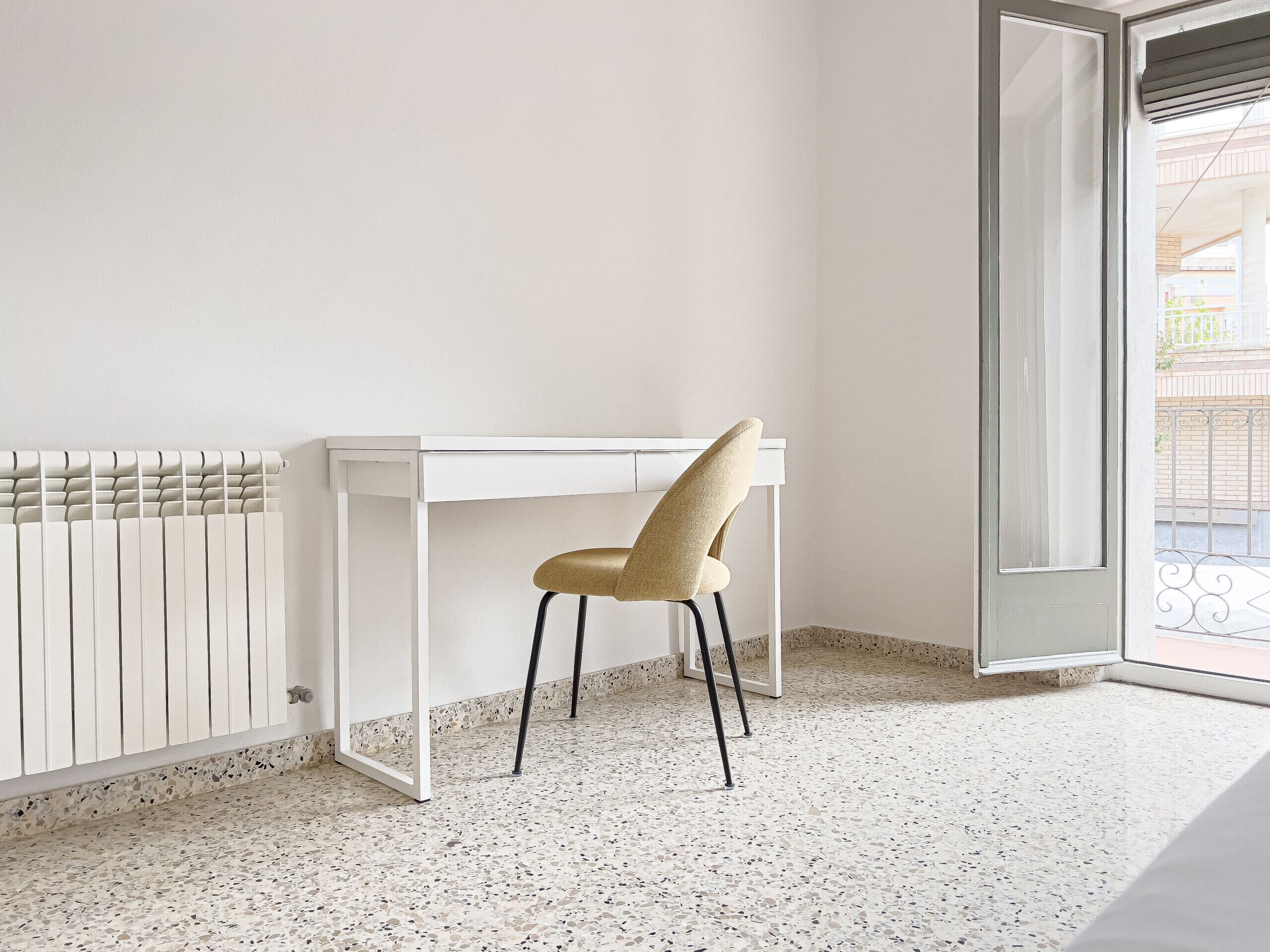 Una habitación con un escritorio blanco, una silla, un radiador y una puerta que da a un balcón exterior.