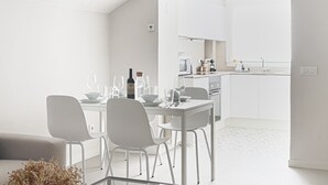 Un comedor moderno y minimalista con una mesa y sillas blancas, una botella de vino y copas. Se ve una cocina al fondo con gabinetes y electrodomésticos blancos.
