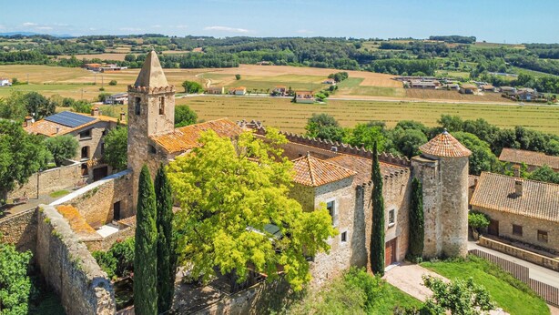 Un edificio de piedra histórico con una torre, rodeado de exuberante vegetación y campos.