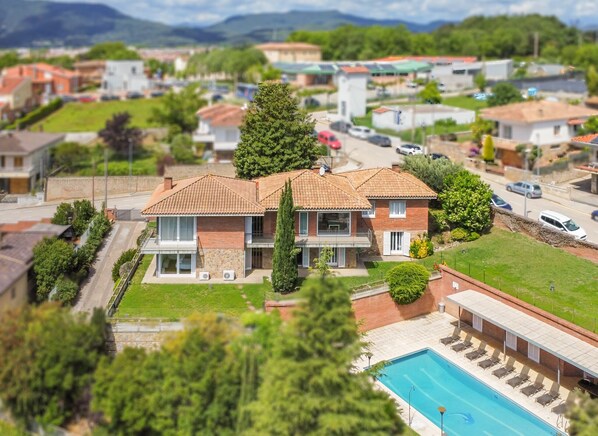 A two-story house with a swimming pool, surrounded by greenery and other houses.