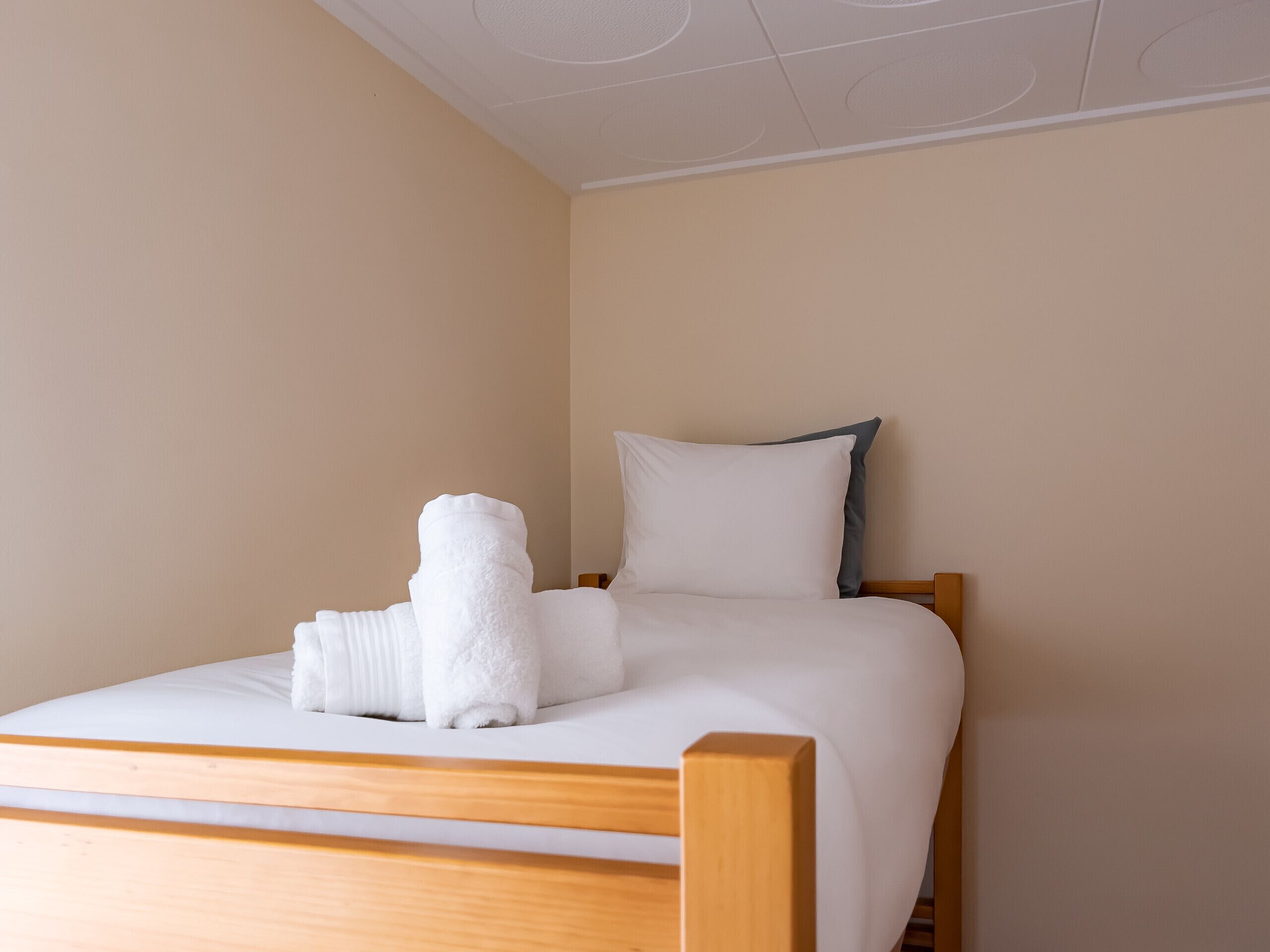 Un lit bien fait, avec une literie blanche et un oreiller gris, une tête de lit en bois et une serviette blanche posée sur le lit.