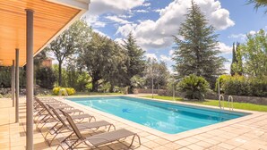 A rectangular swimming pool with a tiled deck, surrounded by lounge chairs and a covered area.