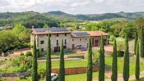 A rustic stone building with solar panels, surrounded by well-maintained gardens and tall cypress trees.