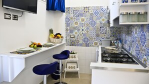 Une petite cuisine équipée d’une cuisinière à gaz, d’un panier de fruits et d’une chaise bleue.
