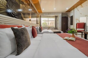 A hotel room with a large bed, a small table with a vase of flowers, a window with a view, and a decorative wall. - Hotel The Amar Grand (Dehradun)
