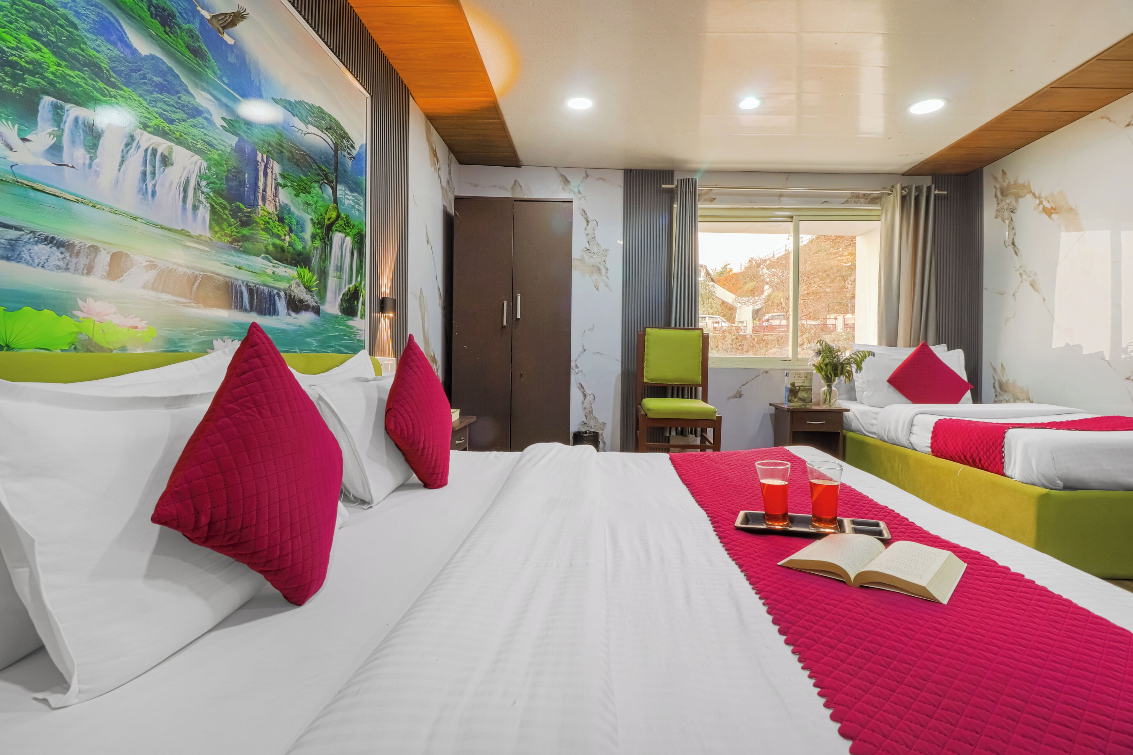A hotel room with a large mural of a waterfall, a bed with white bedding and red pillows, a green chair, and a window with a view of buildings.