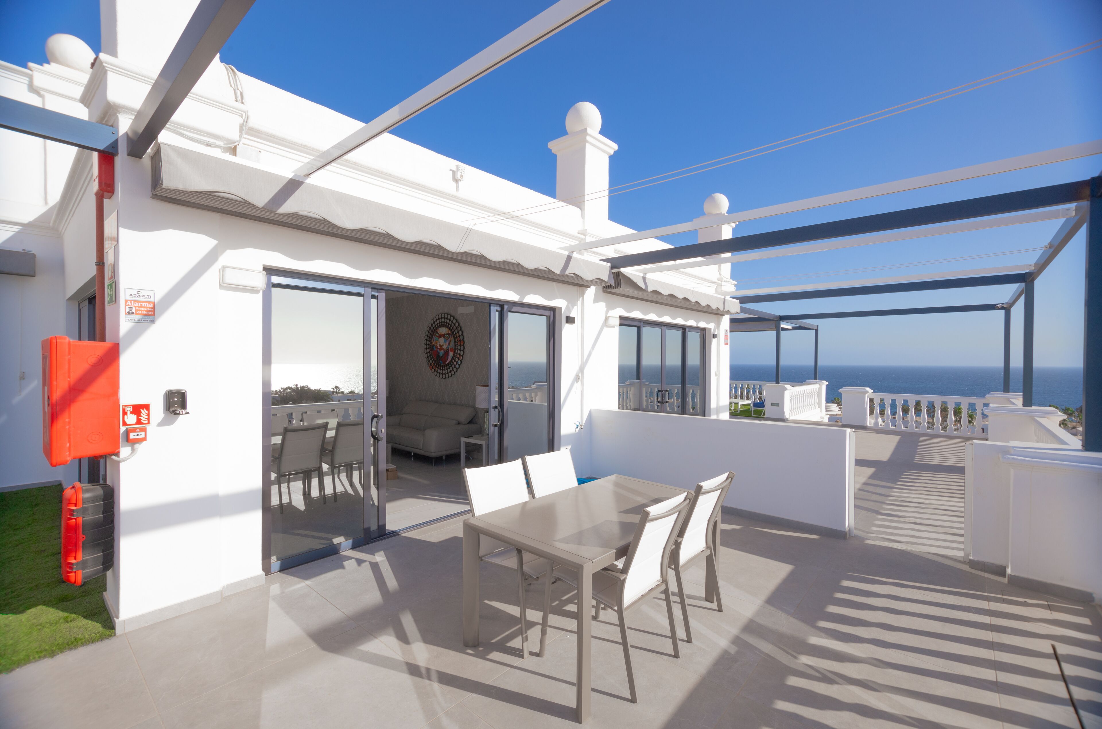 A modern outdoor patio with a dining table and chairs, a glass door leading to a living area, and a view of the ocean.
