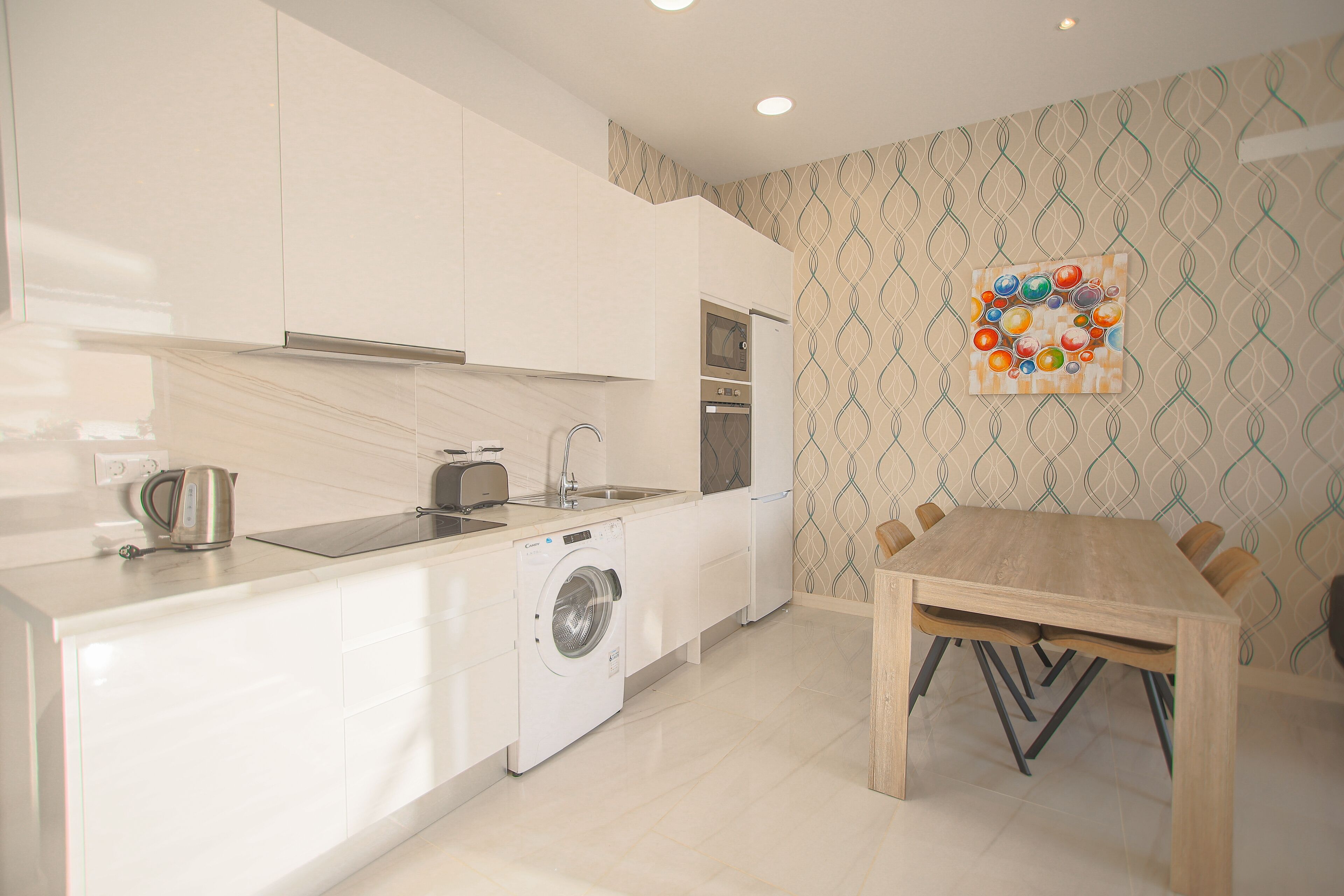 A modern kitchen with white cabinetry, a built-in washing machine, a wooden dining table, and a patterned wall.